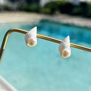 May include: A pair of white seashell earrings with a pearl accent. The earrings are displayed on a gold-colored metal bar, with a blurred background of a swimming pool and greenery.