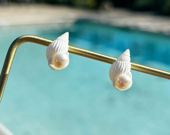 Könnte beinhalten: Ein Paar weiße Muschelohrringe mit Perlenakzent. Die Ohrringe werden auf einem goldfarbenen Metallstab präsentiert, mit einem verschwommenen Hintergrund eines Schwimmbeckens und Grün.