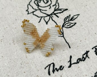 Könnte beinhalten: Ein bestickter Schmetterlings-Anstecker mit weißen und beigefarbenen Flügeln und goldenen Akzenten. Der Schmetterling befindet sich auf cremefarbenem Stoff mit einer schwarzen Rosenillustration und dem Text "The Last Sonnet".