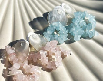 Könnte beinhalten: Zwei Paar Edelsteinohrringe. Ein Paar hat hellblaue Steine, das andere rosa. Jeder Ohrring hat eine kreisförmige, irisierende Rückseite. Die Ohrringe werden auf einer weißen, muschelförmigen Oberfläche präsentiert.