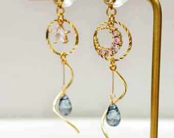 Könnte beinhalten: Goldfarbene Ohrringe mit Stern- und Halbmond-Design. Jeder Ohrring hat eine blaue, tropfenförmige Perle und einen zarten Spiralakzent. Die Ohrringe werden auf einem goldenen Ständer präsentiert.