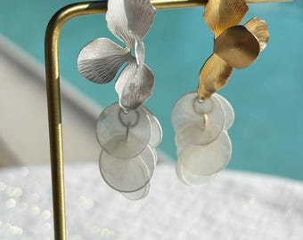 Könnte beinhalten: Ein Paar Ohrringe mit floralem Design. Ein Ohrring hat eine silberfarbene Blütenform und eine Kaskade aus kleinen, runden, weißen Scheiben. Der andere hat eine goldfarbene Blütenform und ähnliche weiße Scheiben.