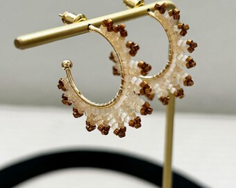 Könnte beinhalten: Goldfarbene Creolen mit Perlenmuster. Die Ohrringe haben eine Halbkreisform mit kleinen, durchsichtigen Perlen und braunen Akzenten. Die Ohrringe werden auf einem goldenen Ohrringständer präsentiert.