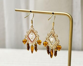Könnte beinhalten: Ein Paar goldfarbene Ohrhänger mit einem rautenförmigen Filigranrahmen, einer zentralen weißen Perle sowie bernsteinfarbenen Glasperlen und braunen facettierten Perlen, die an der Unterseite der Metallstruktur hängen.