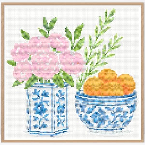 以下が含まれることがあります： クロスステッチのアートワークで、静物画が描かれています。 ピンクの花が青と白の花瓶に入っており、オレンジのボウルが隣にあります。 緑の葉と木製のフレームがデザインを完成させています。