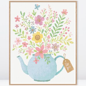以下が含まれることがあります： 額縁に入ったクロスステッチのアート作品で、色とりどりの花で溢れる水色のティーポットが描かれています。花にはピンク、黄色、青の花と緑の葉が含まれています。小さなタグがティーポットのハンドルからぶら下がっています。