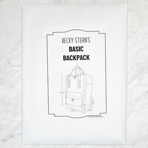 以下が含まれることがあります： 白い紙のパッケージに、バックパックの白黒イラストが描かれています。パッケージのテキストは「Becky Stern's Basic Backpack」と書かれています。バックパックのデザインには、cm単位の寸法が含まれています。