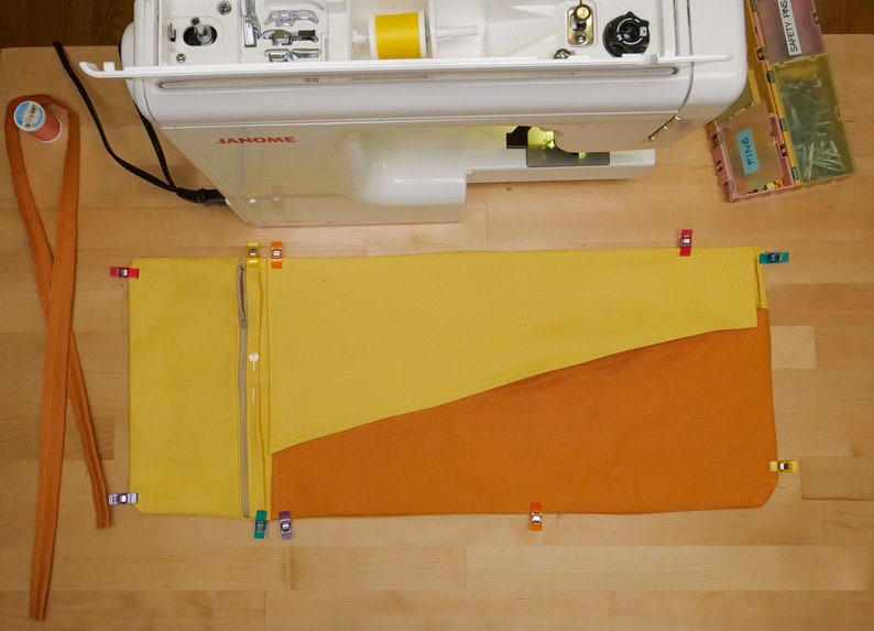 May include: A yellow and orange fabric pouch with a zipper is pinned to a wooden surface. The pouch is being prepared for sewing on a white sewing machine.