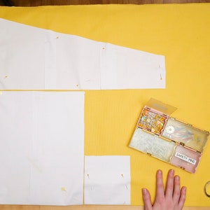 May include: A yellow fabric surface with white paper pattern pieces pinned to it. A small box of safety pins and a pair of scissors are on the surface.
