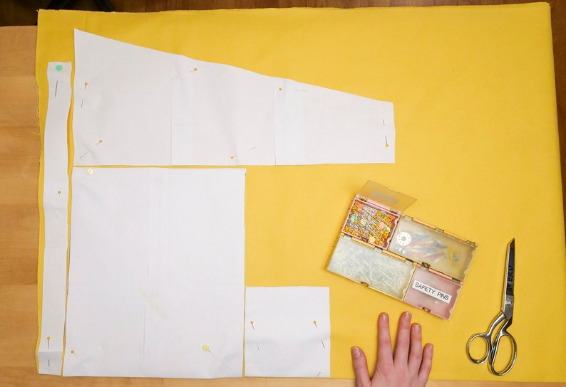 May include: A yellow fabric surface with white paper pattern pieces pinned to it. A small box of safety pins and a pair of scissors are on the surface.