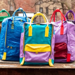 Può includere: Cinque zaini colorati con cinturini e dettagli di diversi colori. Gli zaini sono allineati in fila su una superficie di legno.