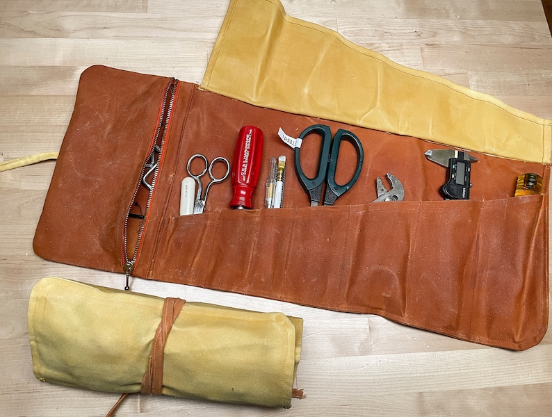 May include: A brown leather tool roll with a zipper closure and a yellow canvas flap. The roll is unrolled and contains a variety of tools, including a pair of scissors, a screwdriver, a wrench, a caliper, and a pencil.