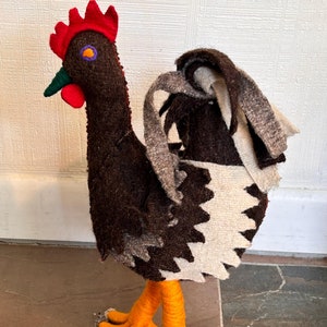 May include: A handcrafted rooster figurine with a brown body, a red comb and wattle, and orange legs. The rooster's tail features a black and white patterned design. The bird's beak is green, and the eye is purple.