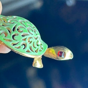 May include: A green and gold turtle brooch with a red rhinestone eye. The turtle's shell is made of openwork metal with a delicate floral design.