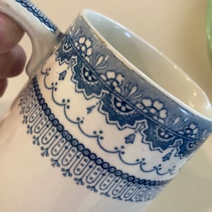 May include: White ceramic mug with a blue floral and scrollwork design around the rim. The mug has a curved handle and a simple, elegant shape. The blue pattern is detailed and intricate, adding a touch of classic style.