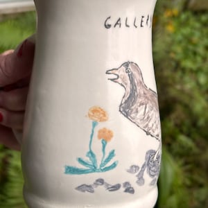 May include: White ceramic mug with a hand-painted design of a bird, flowers, and rocks. The mug has the word "GALLERY" written on the side.