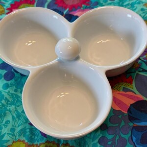 May include: A white ceramic serving dish featuring three interconnected circular bowls arranged in a clover-like pattern. A small, smooth, spherical knob is positioned in the center where the three bowls meet, providing a central point of connection.
