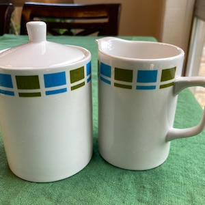 May include: White ceramic sugar bowl with lid and creamer. Both pieces have a band of blue and green squares around the top. The sugar bowl is cylindrical, and the creamer has a handle.