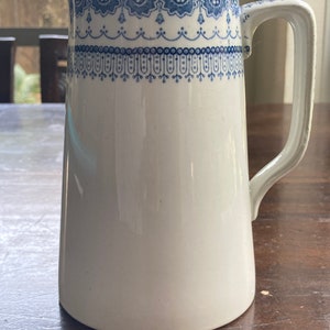 May include: White ceramic pitcher with a blue floral design around the rim and handle. The pitcher has a tall, slender shape and a curved handle. The blue design features a repeating pattern of flowers and leaves.