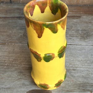 May include: A handmade yellow ceramic tumbler with brown and green accents. The rim features a brown drip design, and green and brown accents encircle the body. The tumbler has a cylindrical shape and a glossy finish.