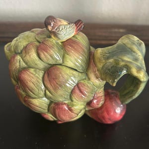 May include: A ceramic artichoke-shaped container with a small bird figurine perched on top. The container is painted in shades of green and red, with a handle and a red bulbous base. The bird has brown, yellow, and red details.