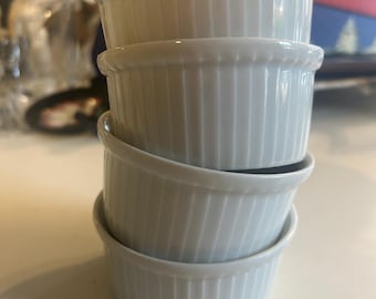 May include: A stack of four white, ribbed ceramic ramekins. Each ramekin has a rounded rim and a slightly flared shape. The ramekins are stacked on top of each other, creating a visually appealing arrangement.