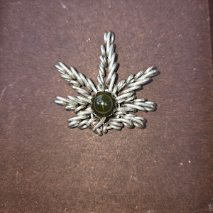 Könnte beinhalten: Ein silberner Anhänger in Blattform mit einem grünen Edelstein in der Mitte.