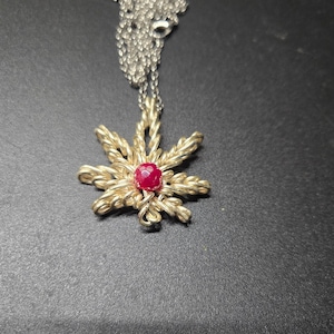 Op de afbeelding: Een gouden ketting met een hanger in de vorm van een gestileerd cannabisblad, met een rode edelsteen in het midden.
