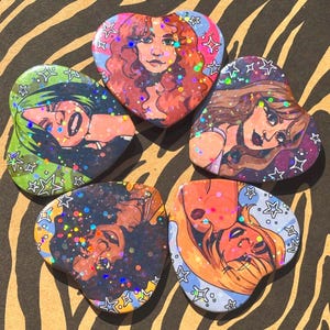 May include: Five heart-shaped pins with illustrations of women. Each pin features a different woman with unique hairstyles and colors. The pins have a glittery, holographic effect and are surrounded by star-shaped borders.