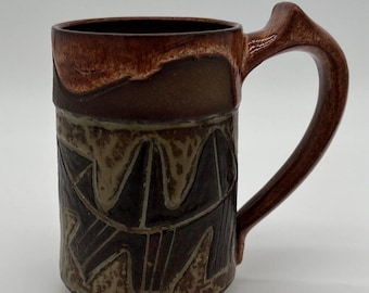 May include: A brown ceramic mug with a geometric design in shades of brown, green, and black. The mug has a rounded body and a handle.