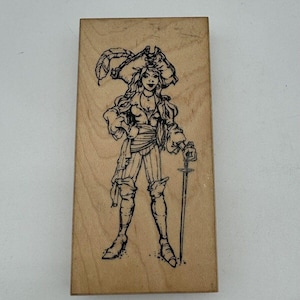 May include: A wooden stamp with a black and white illustration of a pirate with a sword and a feathered hat.