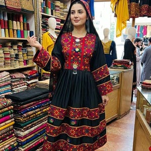 May include: A woman wearing a black dress with red and gold embroidery. The dress has long sleeves and a full skirt. The woman is in a shop filled with colorful fabrics and clothing.