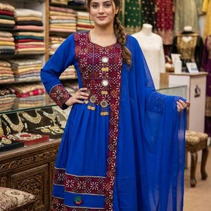 May include: A woman wearing a vibrant blue dress with intricate embroidery and embellishments. The dress features a detailed pattern in burgundy and gold, with matching accents on the sleeves and hem. A sheer blue scarf drapes elegantly.
