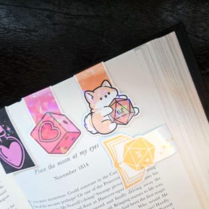 May include: Bookmarks with cute illustrations of a potion bottle, a heart-shaped die, a Shiba Inu holding a D20 die, and a D20 die on a stack of books. The bookmarks are placed in an open book with the text "Place the moon at my eyes November 1814".
