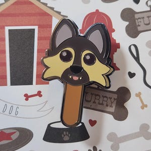 May include: A brown and yellow dog head enamel pin with a black outline. The dog is smiling with its tongue sticking out. The pin is attached to a brown stick and a black metal base.