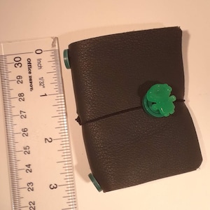 Peut inclure: Un petit carnet en cuir gris foncé avec un fermoir en plastique vert et un bouton en plastique vert. Le carnet est fermé et mesure environ 7,6 cm de long.