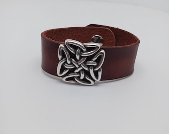May include: A brown leather bracelet with a silver Celtic knot design. The bracelet is adjustable and has a buckle closure.
