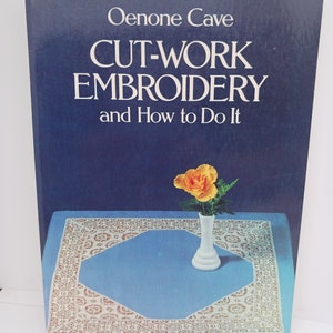 May include: A book titled "Cut-Work Embroidery and How to Do It" by Oenone Cave. The cover features a blue background with a white vase holding a single yellow flower and a white doily on a blue surface.