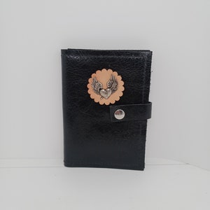 May include: Black leather wallet with a circular cutout featuring a winged heart design. The wallet has a snap closure and a small strap.