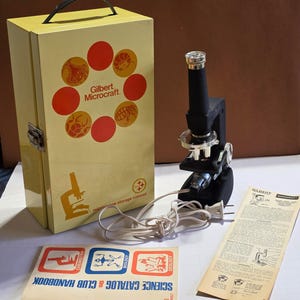 May include: A vintage Gilbert Microcraft microscope set featuring a yellow metal storage cabinet with a black handle, a black electric microscope with a white power cord, a science catalog and club handbook, and an instructional paper sheet.