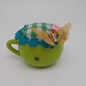 May include: Green ceramic teacup with polka dots and a green and white gingham fabric lid with a gold butterfly and pink flower accent.