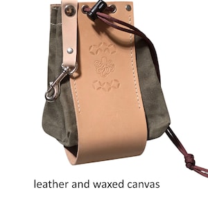 May include: A small, olive green pouch with a drawstring closure, featuring a tan leather accent with an embossed design. The pouch is made of leather and waxed canvas, with a metal clasp and the text "drawstring closure" and "leather and waxed canvas".