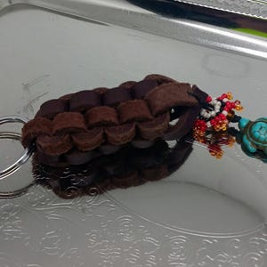 May include: A brown leather keychain with a braided design and a turquoise turtle charm. The keychain also features a beaded charm with red, white, and blue beads.