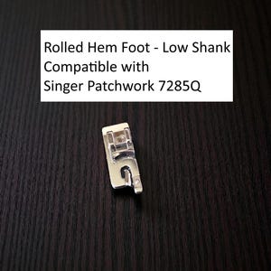 May include: A silver-colored rolled hem foot for sewing machines. The text on the image reads: "Rolled Hem Foot - Low Shank Compatible with Singer Patchwork 7285Q". The sewing machine accessory is on a dark wood grain surface.