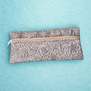 May include: A rectangular, gray pouch with a floral embossed pattern. The pouch has a beige zipper closure and a white zipper pull. The background is a textured, light blue surface.