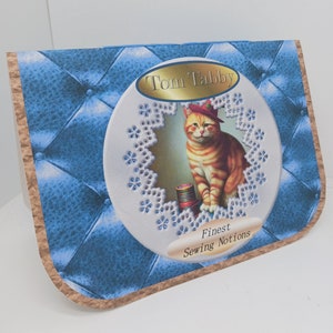 May include: A blue and brown card with a white circle in the center. The circle has a white lace design around the edge and a picture of a cat wearing a red hat inside. The text "Tom Tabby Finest Sewing Notions" is printed on the card.