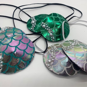 Könnte beinhalten: Vier Augenklappen im Meerjungfrauen-Design mit schillernden Schuppen und Perlenbesatz. Jede Augenklappe ist an einem schwarzen oder weißen Gummiband befestigt.