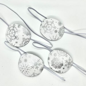 Puede incluir: Cuatro parches para ojos blancos con diseños de copos de nieve plateados y correas elásticas.