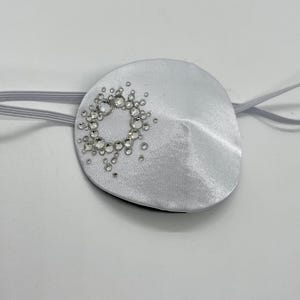 Könnte beinhalten: Eine weiße Augenklappe aus Satin mit einem dekorativen Kreis aus klaren Strasssteinen. Die Augenklappe hat ein dünnes, elastisches Band zur Befestigung am Kopf. Die Strasssteine sind kreisförmig angeordnet.