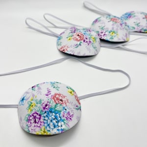 May include: Several floral eye patches with elastic bands are displayed against a white background. The patches feature a white fabric base adorned with a colorful floral pattern in shades of pink, purple, blue, and yellow.